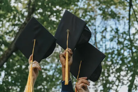 أيادٍ ترفع قبعات تخرج سوداء بشرّابات ذهبية أمام أشجار خضراء